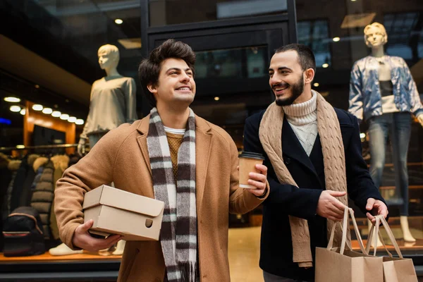 Bearded gay man with shopping bags looking at happy boyfriend with paper cup and shoebox near showcase with mannequins — Stock Photo