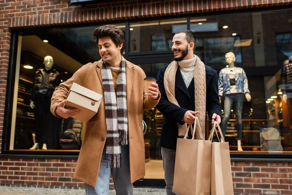 Trendiger schwuler Mann mit Einkaufstaschen schaut fröhlichen Freund mit Schuhkarton und Pappbecher in der Nähe der Vitrine an — Stockfoto