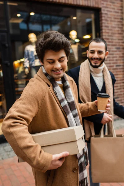 Весёлый гей смотрит на обувную коробку рядом с улыбающимся парнем с пакетом для покупок и бумажной чашкой на размытом витрине — стоковое фото
