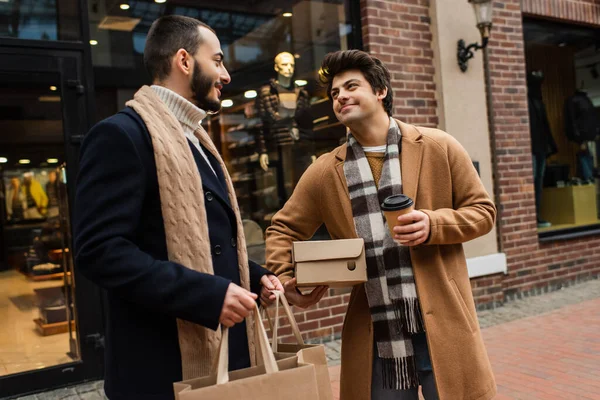 Heureux gay l'homme avec shoebox et à emporter boisson en regardant barbu copain avec shopping sacs près flou magasin — Photo de stock