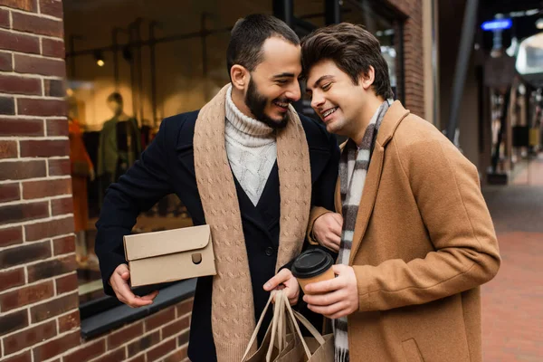 Happy gay couple with purchases and paper cup standing near blurred showcase on city street — Stock Photo