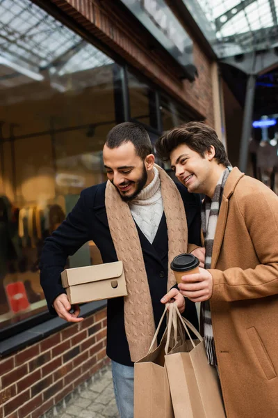 Sorrindo gay homem com café para ir inclinado no barbudo namorado com shoebox e compras sacos perto de vitrine no rua — Fotografia de Stock