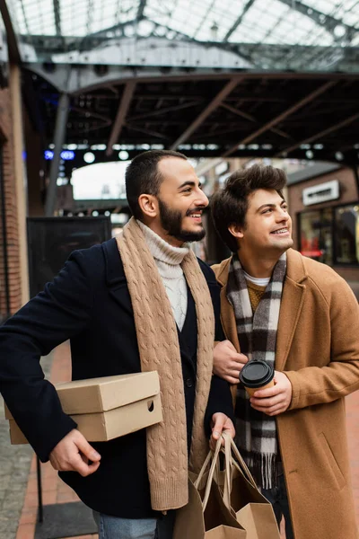 Moda gay casal com compras e café para ir sorrindo e olhando para longe na rua turva — Fotografia de Stock