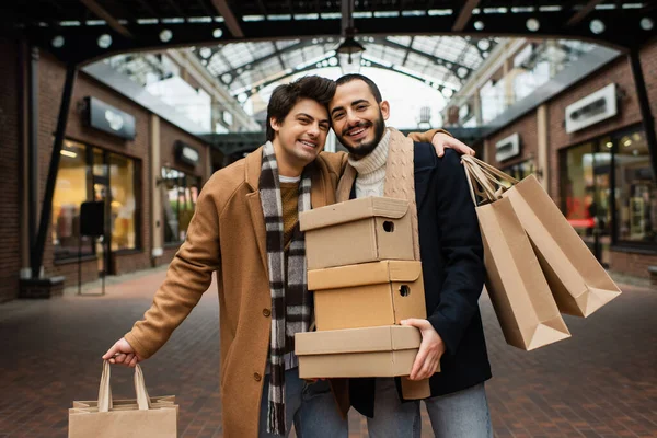 Pleased gay man with shopping bags hugging bearded boyfriend with shoeboxes on urban street — Stock Photo