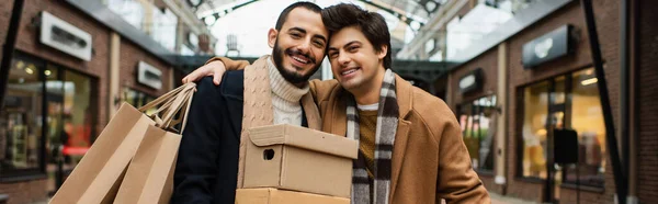 Cheerful man with shopping bags embracing gay partner with shoeboxes near blurred shops on background, banner — Stock Photo