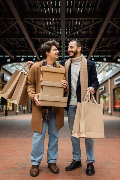 Full length of joyful and stylish gay men with shopping bags and shoeboxes looking at each other on urban street — Stock Photo