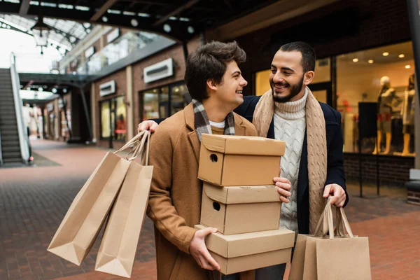 Бородатий гей чоловік з сумками дивиться на щасливого хлопця, що тримає тапочки біля розмитих магазинів на вулиці — стокове фото