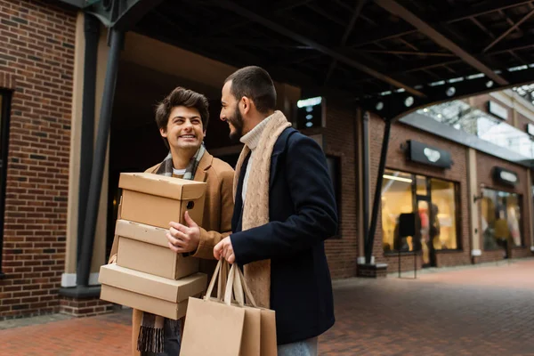 Satisfeito e moderno gay casal com compras sacos e shoeboxes olhando para o outro no urbano rua — Fotografia de Stock