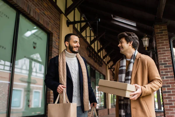 Cheerful man with shoebox looking at bearded boyfriend holding shopping bags near building with showcases — Stock Photo