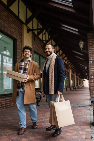 Повна довжина щасливої пари геїв у модному одязі, що стоїть з покупками біля будівлі з магазинами — стокове фото