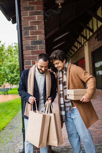Cheerful gay men in stylish clothes looking into shopping bags near building on city street — Stock Photo