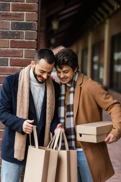 Sonriente pareja homosexual en elegante traje mirando en bolsas de compras cerca de la columna de ladrillo en la calle de la ciudad - foto de stock