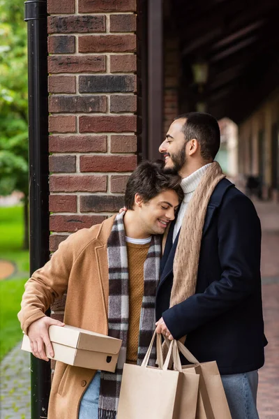 Felice gay uomo con bretelle e chiuso gli occhi tenendo shoebox mentre appoggiato su barbuto fidanzato con shopping bags — Foto stock