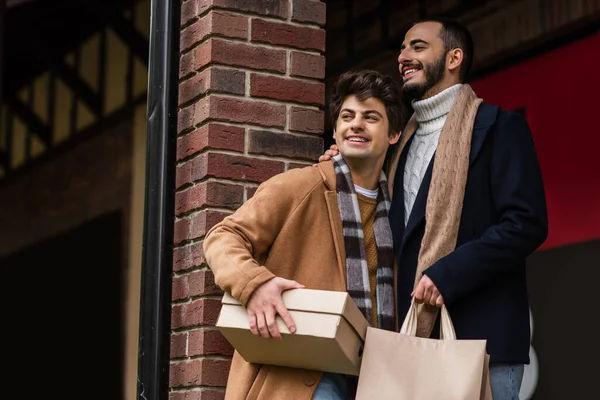 Joyeux et élégant couple gay avec des sacs à provisions et boîte à chaussures regardant loin près de colonne de brique — Photo de stock