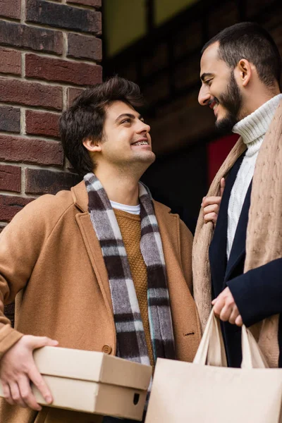 Joyful and trendy gay couple with purchases smiling at each other near brick wall outdoors — Stock Photo