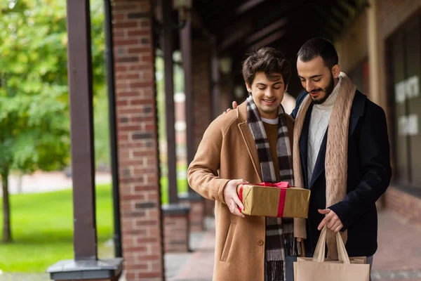 Smiling and trendy gay couple looking at gift box while standing with shopping bags on blurred street — Stock Photo