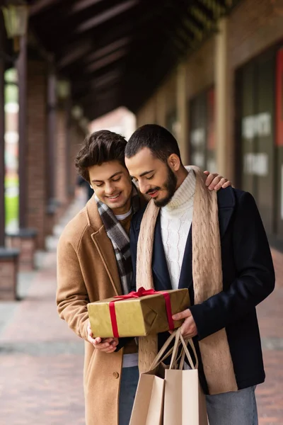 Happy gay man hugging bearded boyfriend holding shopping bags and Christmas presents on blurred street — Stock Photo