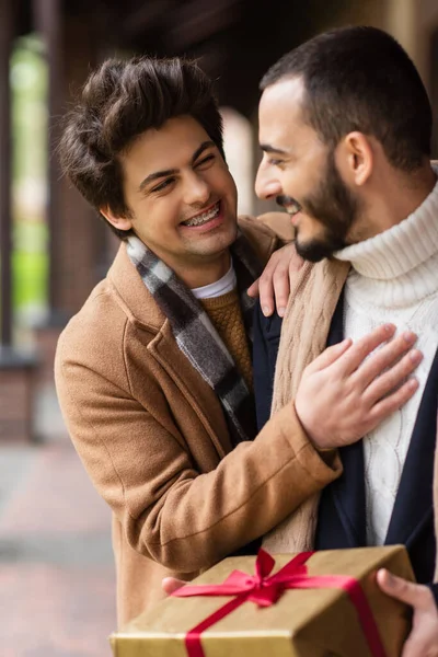 Young and cheerful gay man touching chest of bearded boyfriend holding Christmas present outdoors — Stock Photo