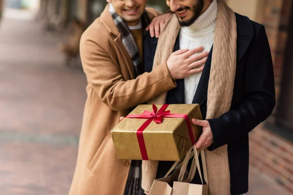 Vista ritagliata di uomo gay barbuto con regalo di Natale e borse della spesa vicino fidanzato sulla strada sfocata — Foto stock