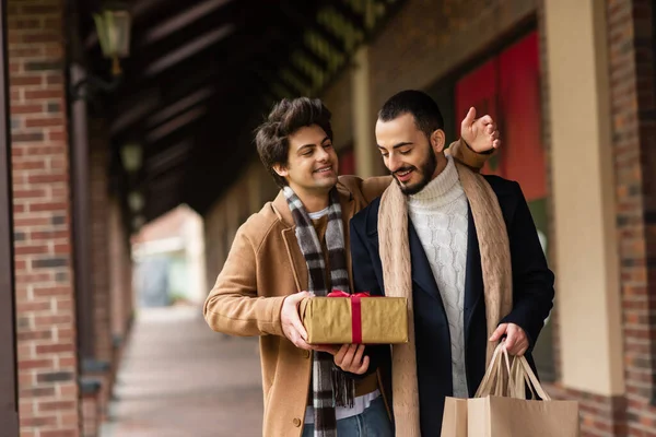 Cheerful gay man presenting gift box to pleased and trendy boyfriend holding shopping bags on city street — Stock Photo