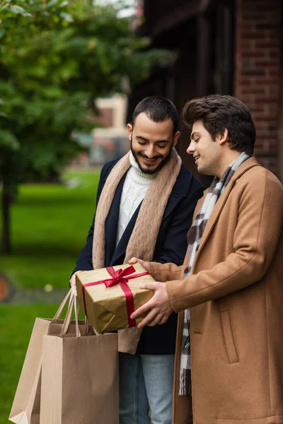 Young man showing Christmas present to bearded boyfriend with shopping bags on blurred street — Stock Photo