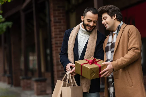 Smiling gay man holding gift box near happy bearded boyfriend with shopping bags on blurred street — Stock Photo