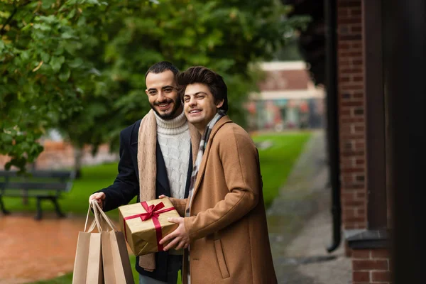 Cheerful and fashionable gay men with christmas present and shopping bags looking at camera outdoors — Stock Photo