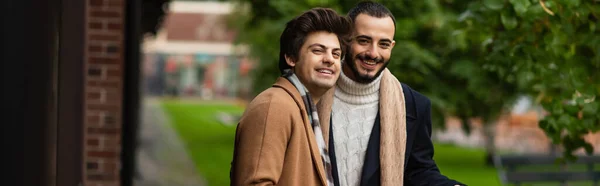 Молода і модна пара геїв у плащах у шарфах посміхається на фотоапараті на вулиці, банер — стокове фото