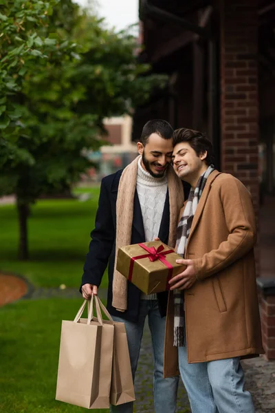 Pleased gay man with closed eyes holding Christmas present while leaning on bearded boyfriend with shopping bags — Stock Photo