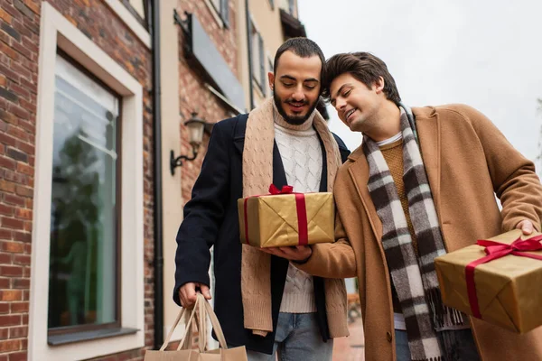 Joven gay hombre celebración regalo cajas cerca feliz y de moda novio con compras bolsas en urbano calle - foto de stock