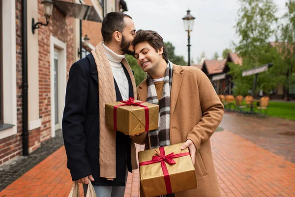 Bearded gay man kissing head of happy boyfriend holding Christmas presents on city street — Stock Photo