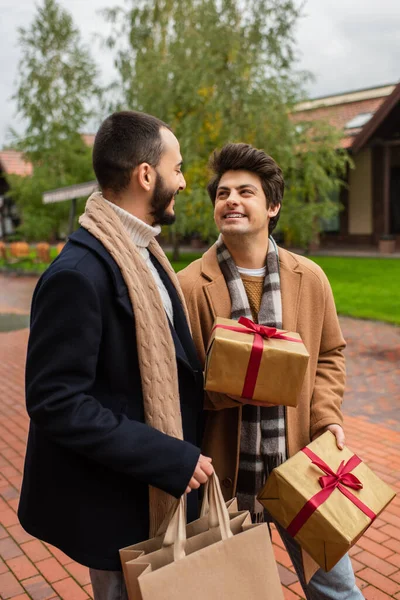 Cheerful gay man holding Christmas presents near trendy boyfriend with shopping bags on blurred street — Stock Photo