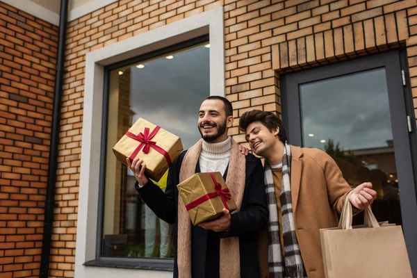 Bearded gay man holding Christmas gift boxes near cheerful boyfriend and shop with showcases — Stock Photo