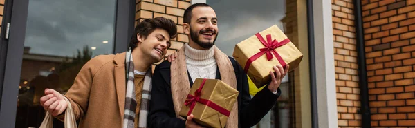 Стильний бородатий чоловік тримає різдвяні подарунки біля веселого хлопця і магазину на вулиці, банер — стокове фото