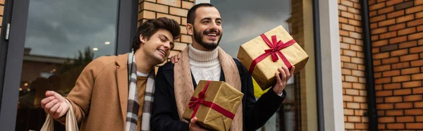 Stylish bearded man holding Christmas presents near cheerful boyfriend and shop on street, banner — Stock Photo