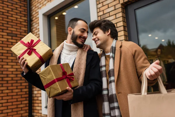 Modisches schwules Paar mit Weihnachtsgeschenken und Einkaufstüte lächelt in der Nähe von Geschäften mit Vitrinen auf der Straße — Stockfoto