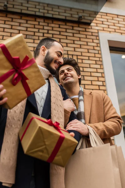 Faible angle de vue des hommes gays joyeux avec des cadeaux de Noël et des sacs à provisions près du bâtiment sur la rue — Photo de stock