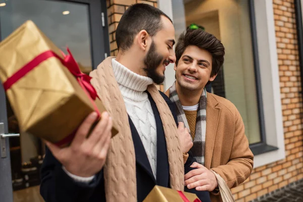 Barbuto gay uomo holding offuscata Natale regalo scatole vicino felice fidanzato con bretelle — Foto stock