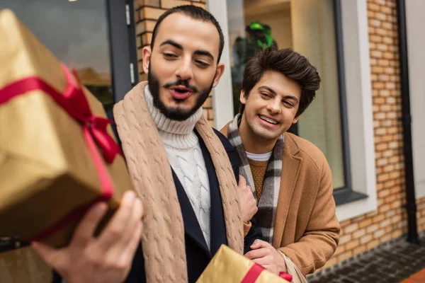 Asombrado gay hombre celebración borrosa Navidad regalo caja cerca alegre novio y tienda en urbano calle - foto de stock