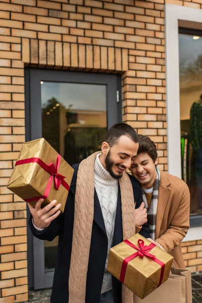 Alegre gay hombre riendo cerca barbudo novio celebración Navidad regalo cajas cerca tienda en calle - foto de stock