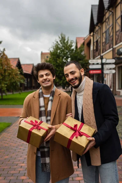 Pareja gay emocionada y de moda con cajas de regalo de Navidad mirando a la cámara en la calle de la ciudad - foto de stock