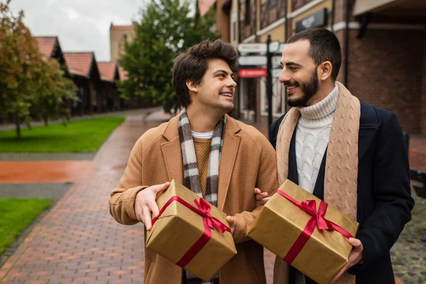 Щаслива гей пара в модних пальто і шарфи тримає подарункові коробки і дивиться один на одного на відкритому повітрі — стокове фото