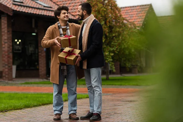 Longitud completa de feliz pareja gay en trajes de moda de pie con cajas de regalo en primer plano borrosa - foto de stock