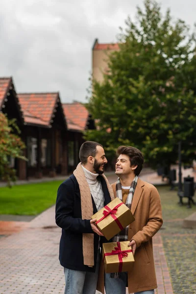 Fashionable gay couple with Christmas presents smiling at each other on street — Stock Photo