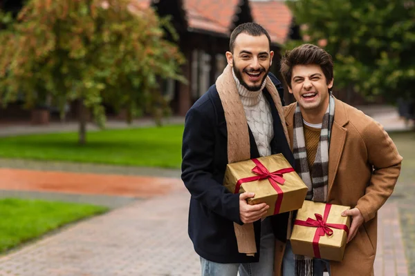 Trendy and cheerful gay men holding gift boxes and laughing at camera on blurred street — Stock Photo