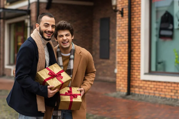 Excited and stylish gay couple with Christmas presents laughing at camera near blurred building — Stock Photo