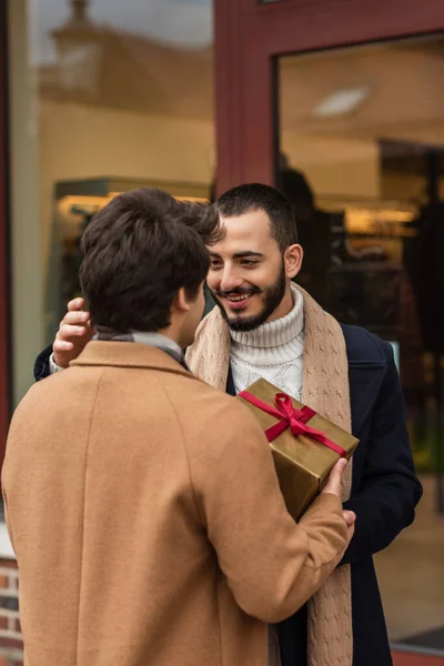 Pleased bearded gay man smiling near boyfriend with Christmas gift box and blurred showcase — Stock Photo