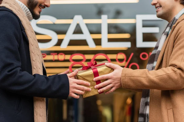 Partial view of smiling gay men holding gift box near blurred showcase with sale lettering — стоковое фото