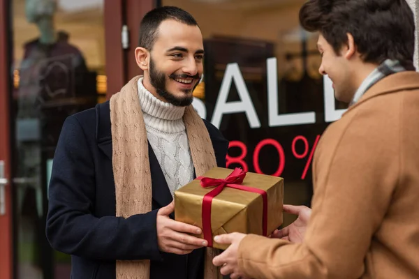 Heureux barbu homme tenant cadeau de Noël près flou copain et vitrine avec vente lettrage — Photo de stock