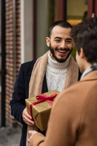 Cheerful bearded gay man holding Christmas present near blurred boyfriend on street — Stock Photo